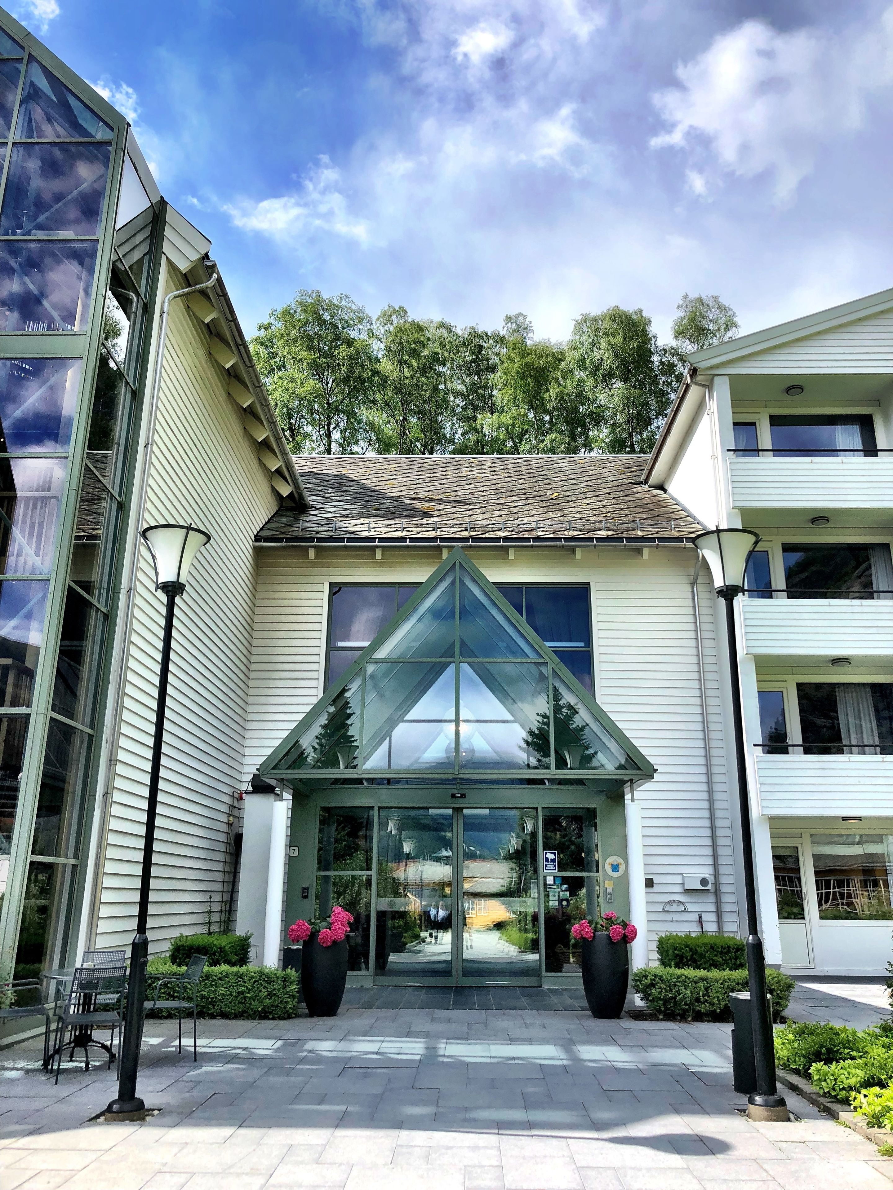 A building with a glass entrance, a tiled roof, and a landscaped entrance area with chairs and potted plants.