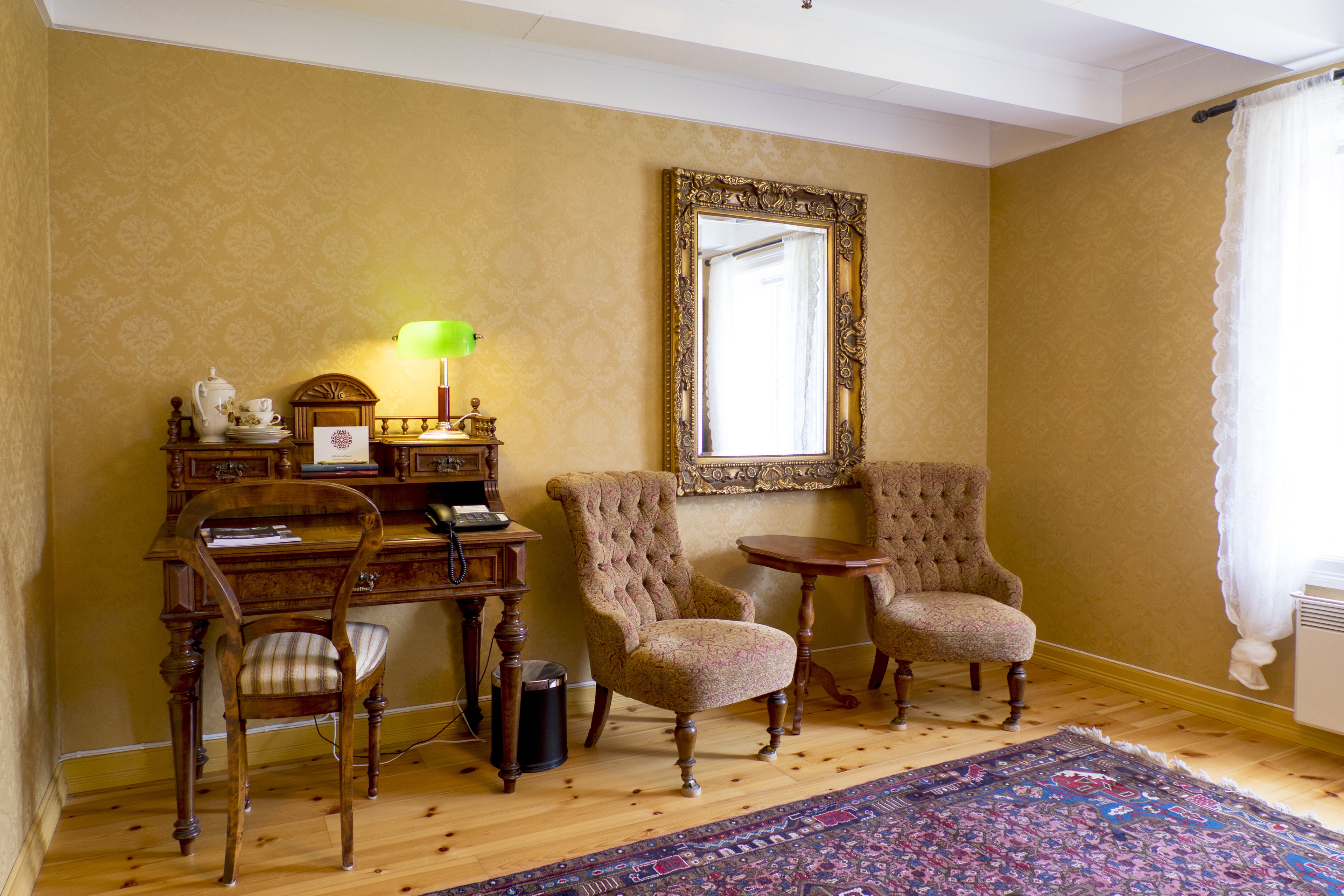 A room with two upholstered chairs, a wooden desk with a lamp, and a large mirror on the wall.
