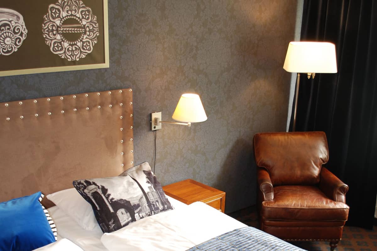 Una habitación de hotel con una cama grande, un sillón de cuero marrón, una mesita de noche con lámpara y una pared decorada con arte.