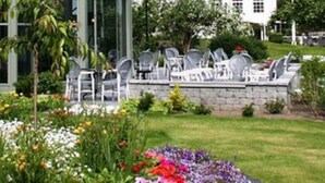 A modern building with a glass facade, surrounded by a well-maintained garden with colorful flowers and a view of a hillside with traditional houses. - Fretheim Hotel (Aurland)