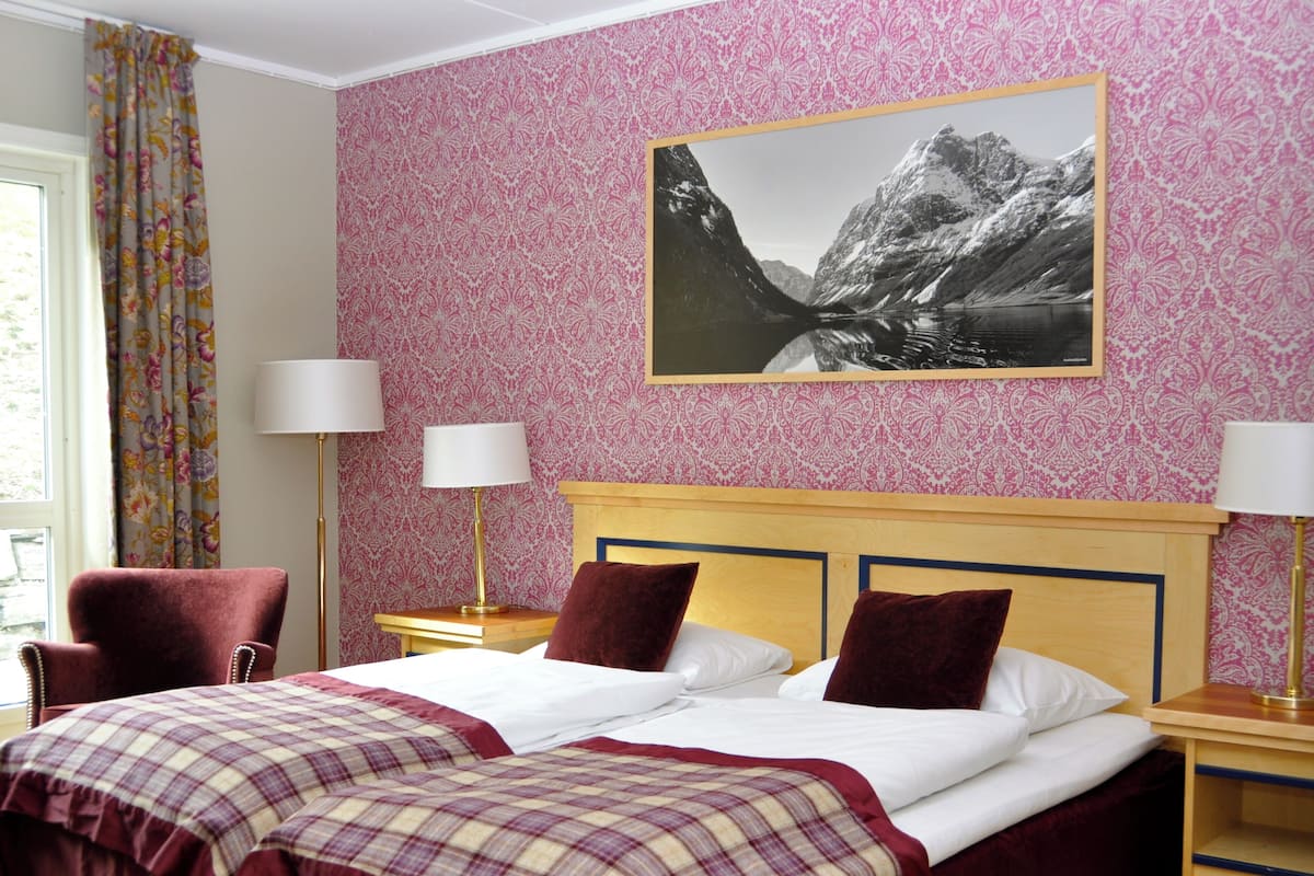 Una habitación de hotel con dos camas, papel tapiz floral, un cuadro de un paisaje de montaña y una silla.