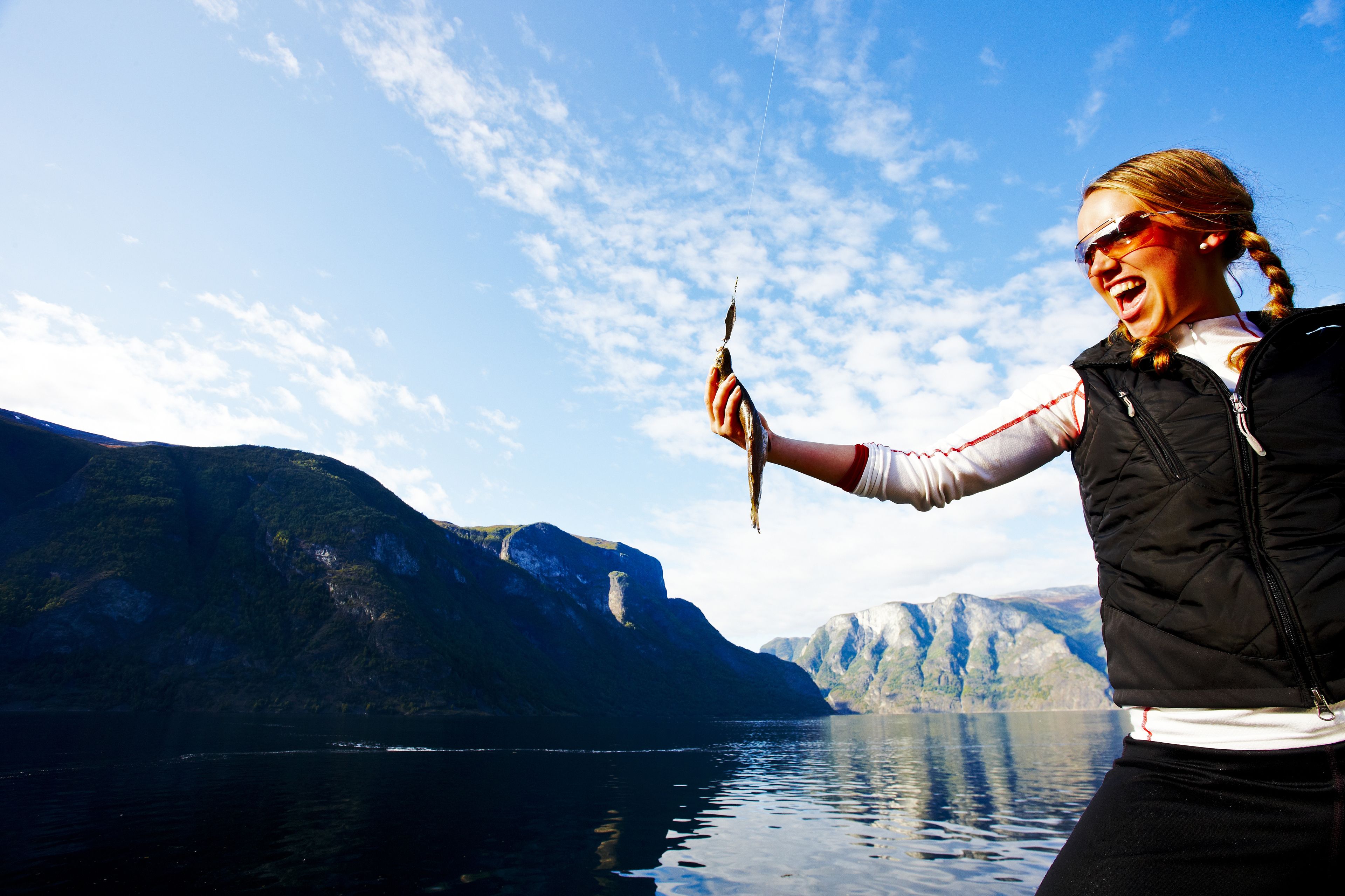 A person holding a fish, standing by a body of water with mountains in the background.