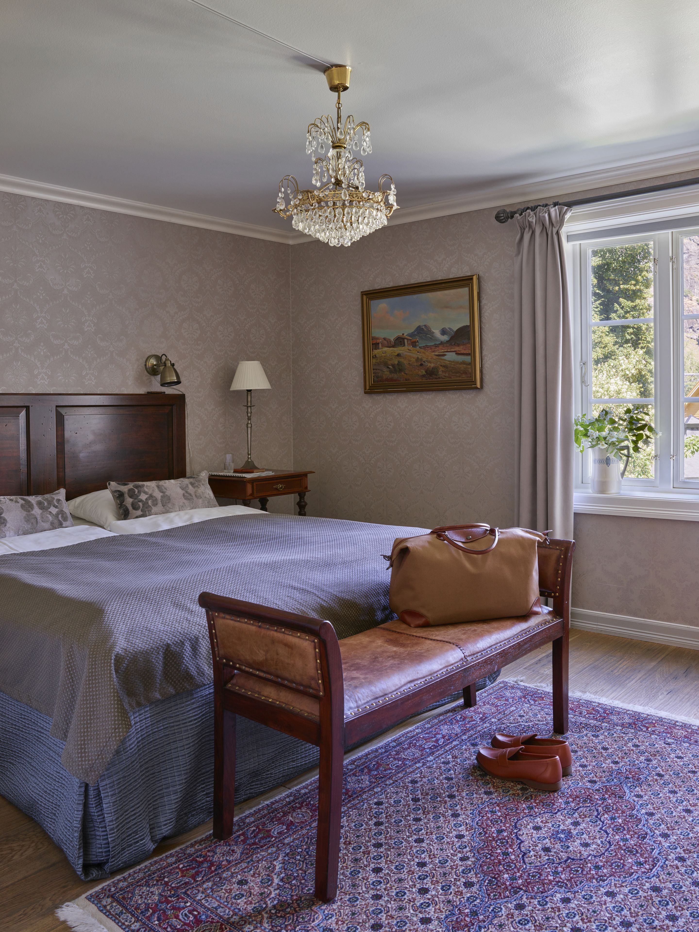 Een slaapkamer met een groot bed, een houten bankje, een kroonluchter, een schilderij en een raam met gordijnen.