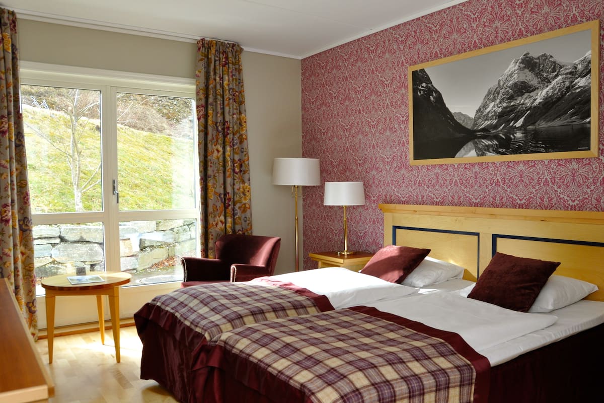 Habitación de hotel con dos camas, una ventana con cortinas, una silla, una mesa y una lámpara.