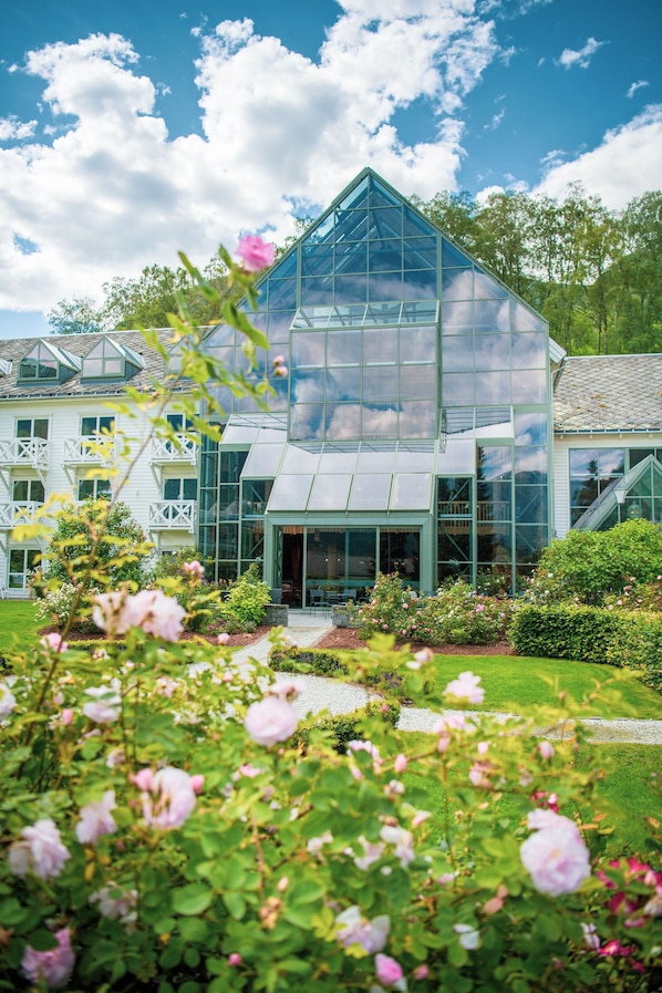 A modern glass greenhouse structure with a triangular roof, surrounded by lush greenery and pink flowers.