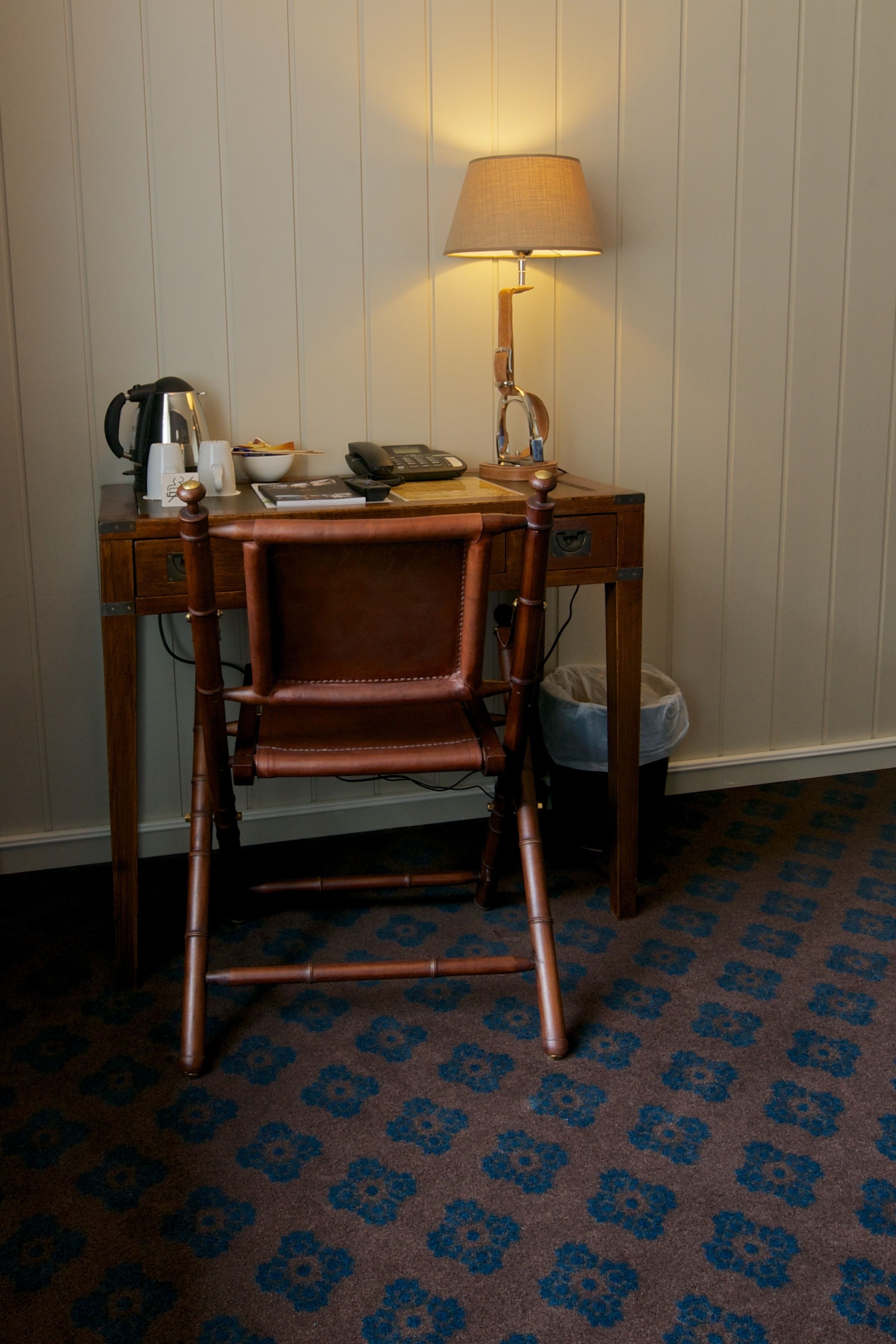 Een houten bureau met een lamp, een stoel en een prullenbak.
