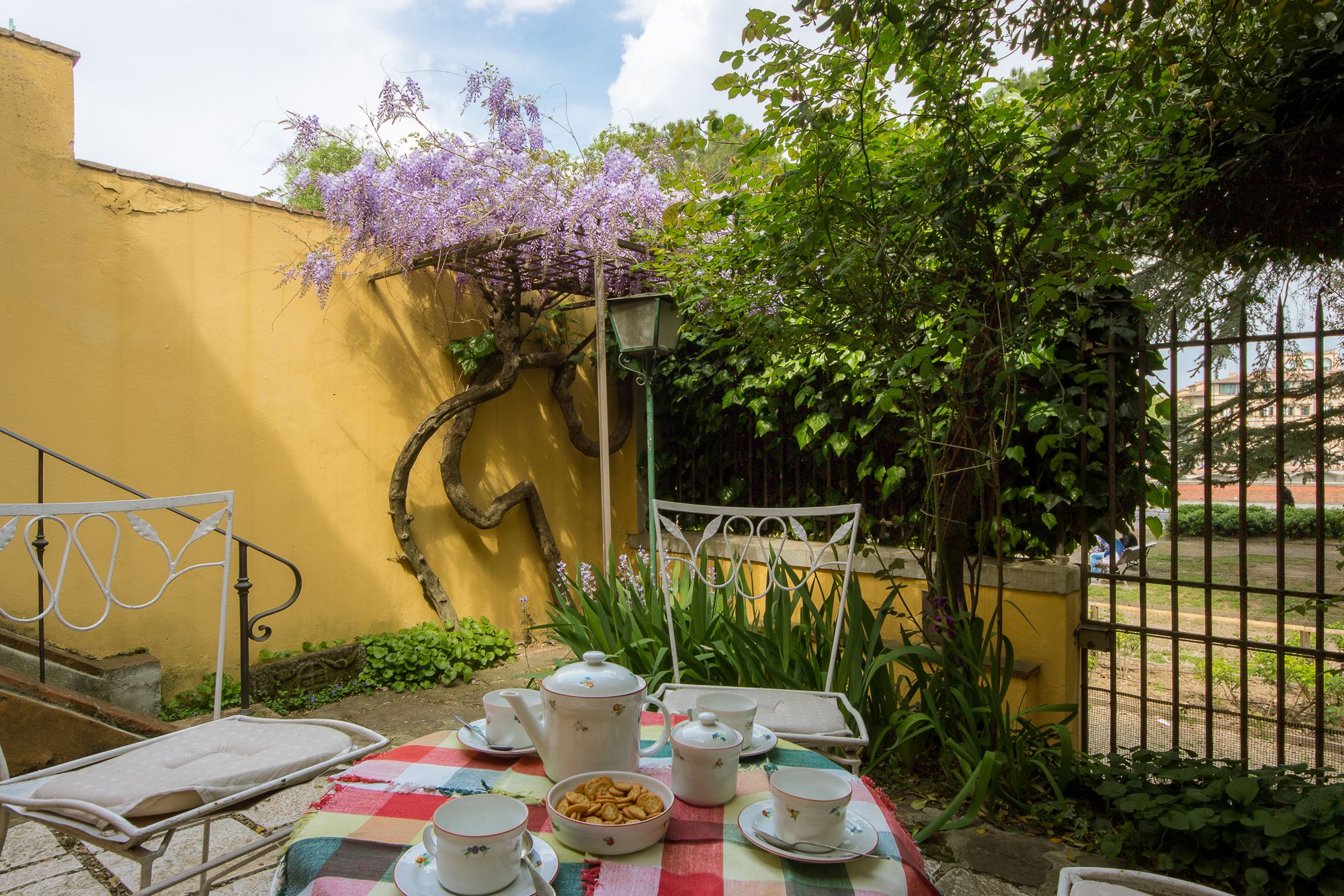 Un patio con una mesa dispuesta para té, rodeado de paredes amarillas y flores púrpuras.