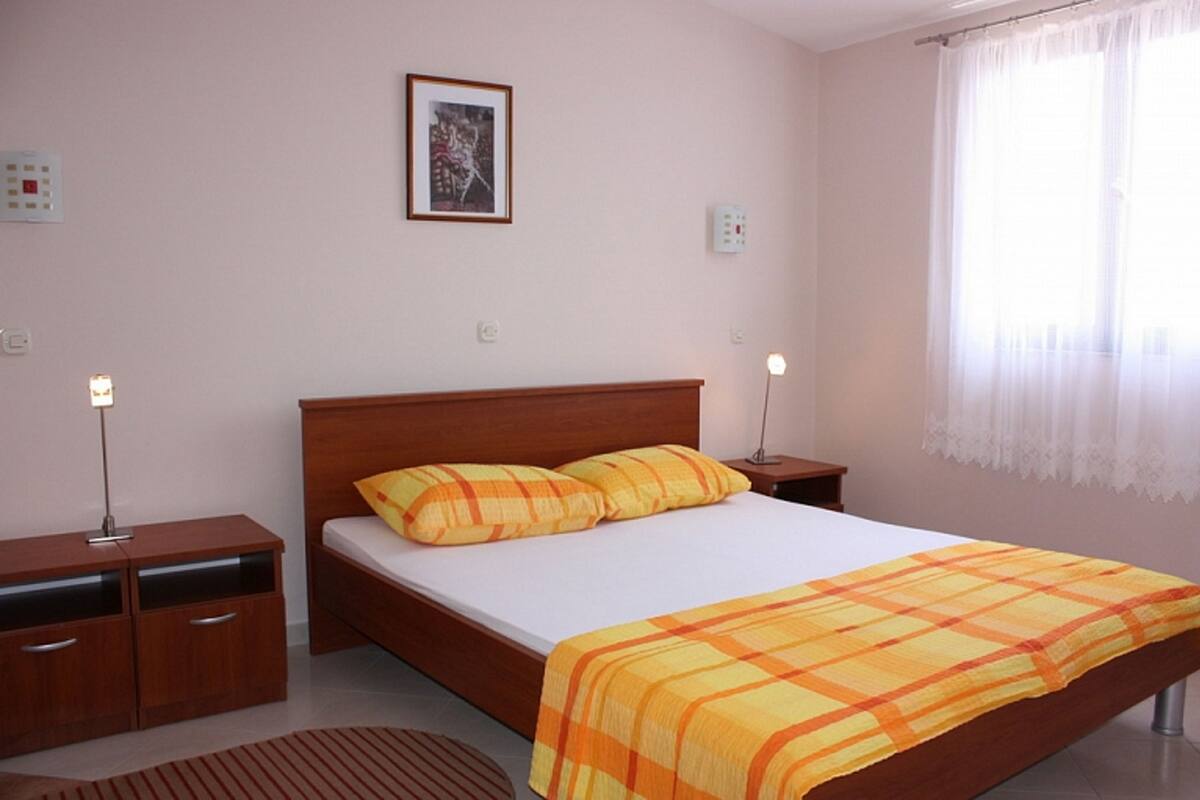 A bedroom with a wooden bed, two bedside tables, a nightstand, a framed picture on the wall, and a window with sheer curtains.