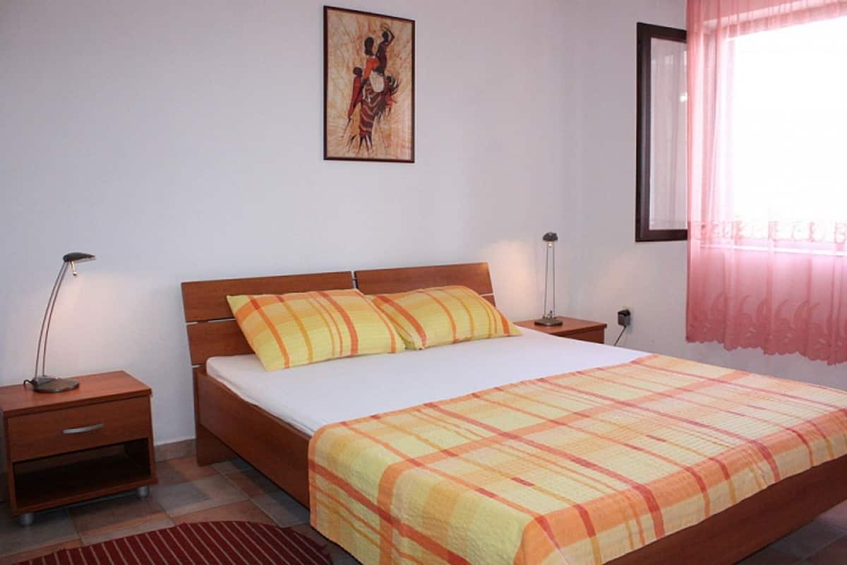 A bedroom with a wooden bed, two bedside tables, a nightstand, a window with pink curtains, and a wall-mounted picture.