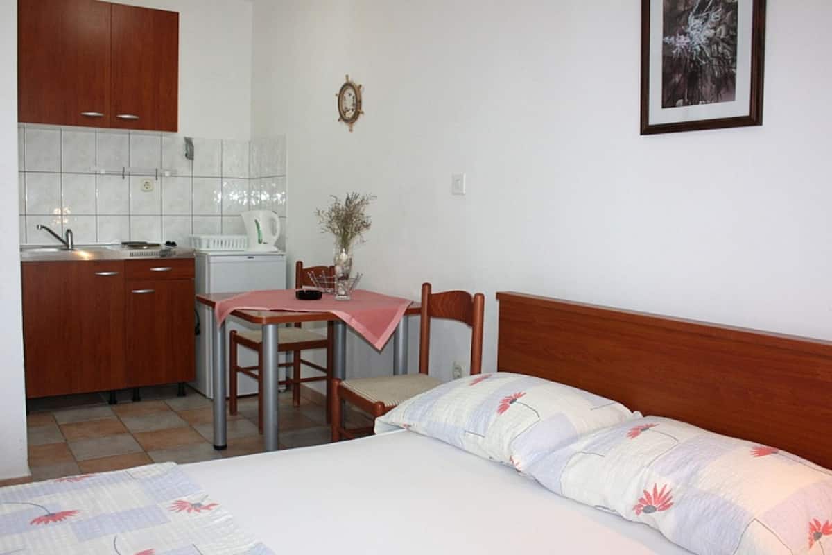 A compact room with a bed, a small table with chairs, a kitchenette with cabinets and a sink, and a framed picture on the wall.