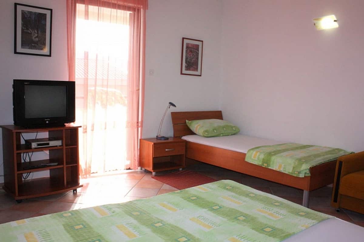 A bedroom with a bed, a TV on a stand, a bedside table, and a window with curtains.