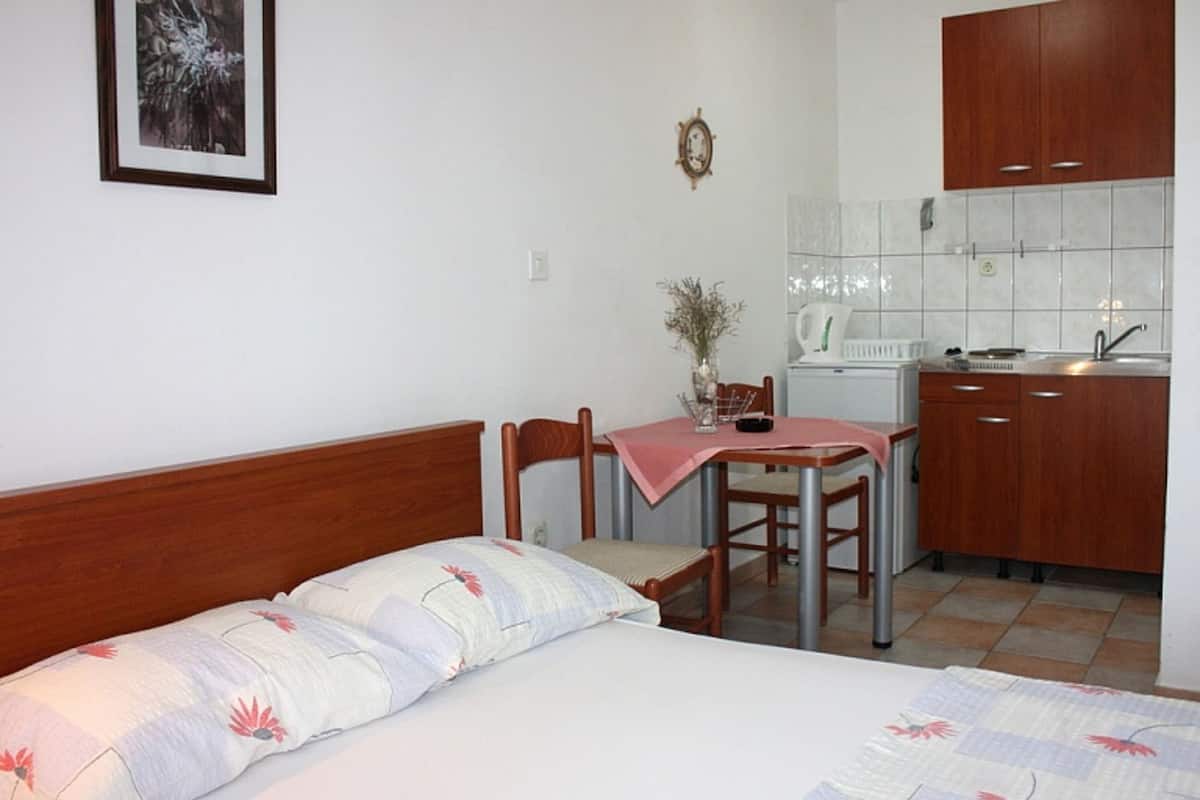 A bedroom with a bed, a bedside table, a chair, a small table with a vase, and a kitchenette with cabinets and a sink.