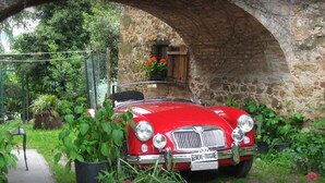 Un coche clásico rojo estacionado bajo un arco de piedra con follaje y una ventana de madera arriba.