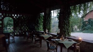 Un área de comedor al aire libre con mesas y sillas, cubierta por una pérgola con enredaderas, y vistas a un jardín y un edificio al fondo.