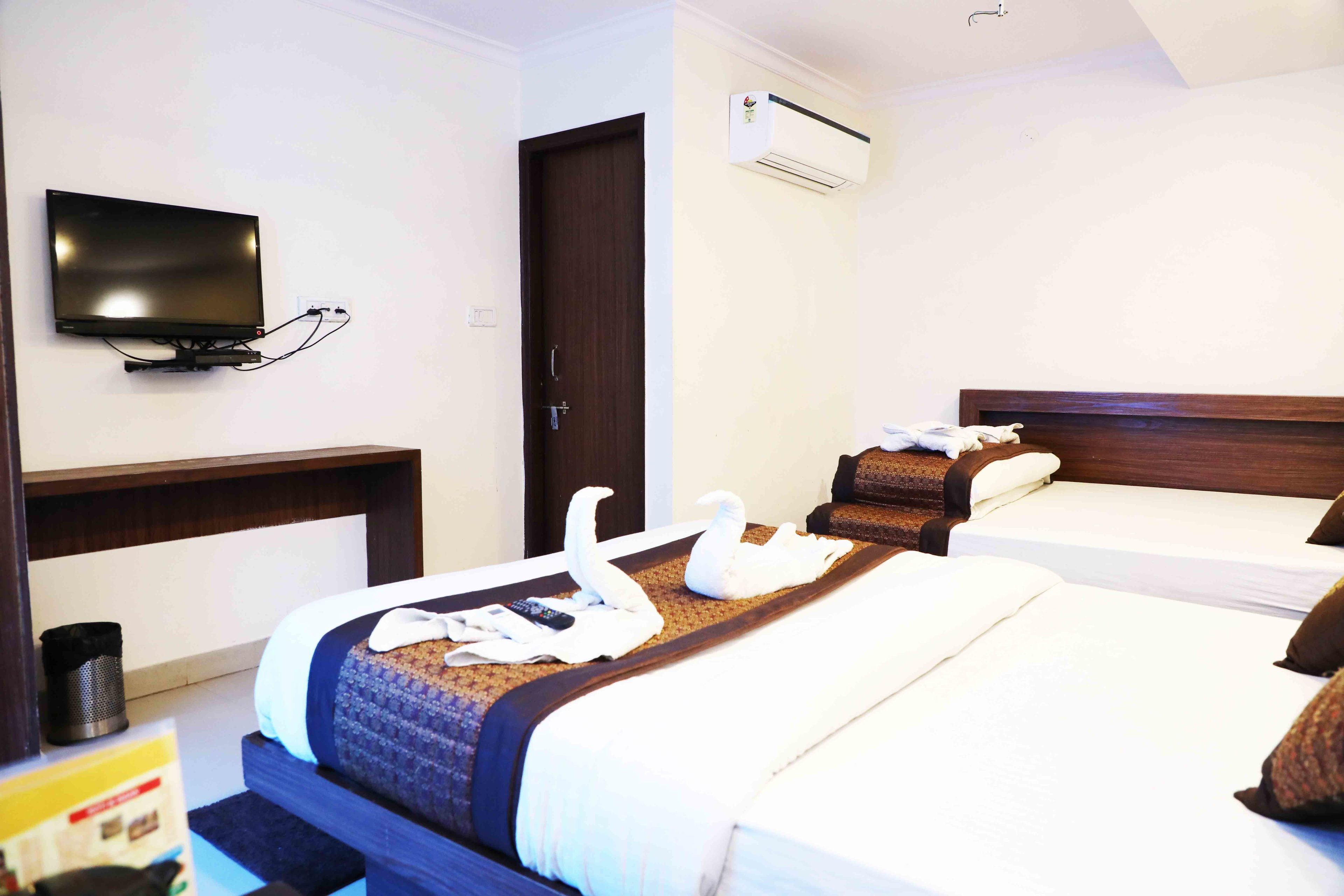 A hotel room with a TV mounted on the wall, a bed with a wooden headboard, a bedside table, and an air conditioning unit on the wall.