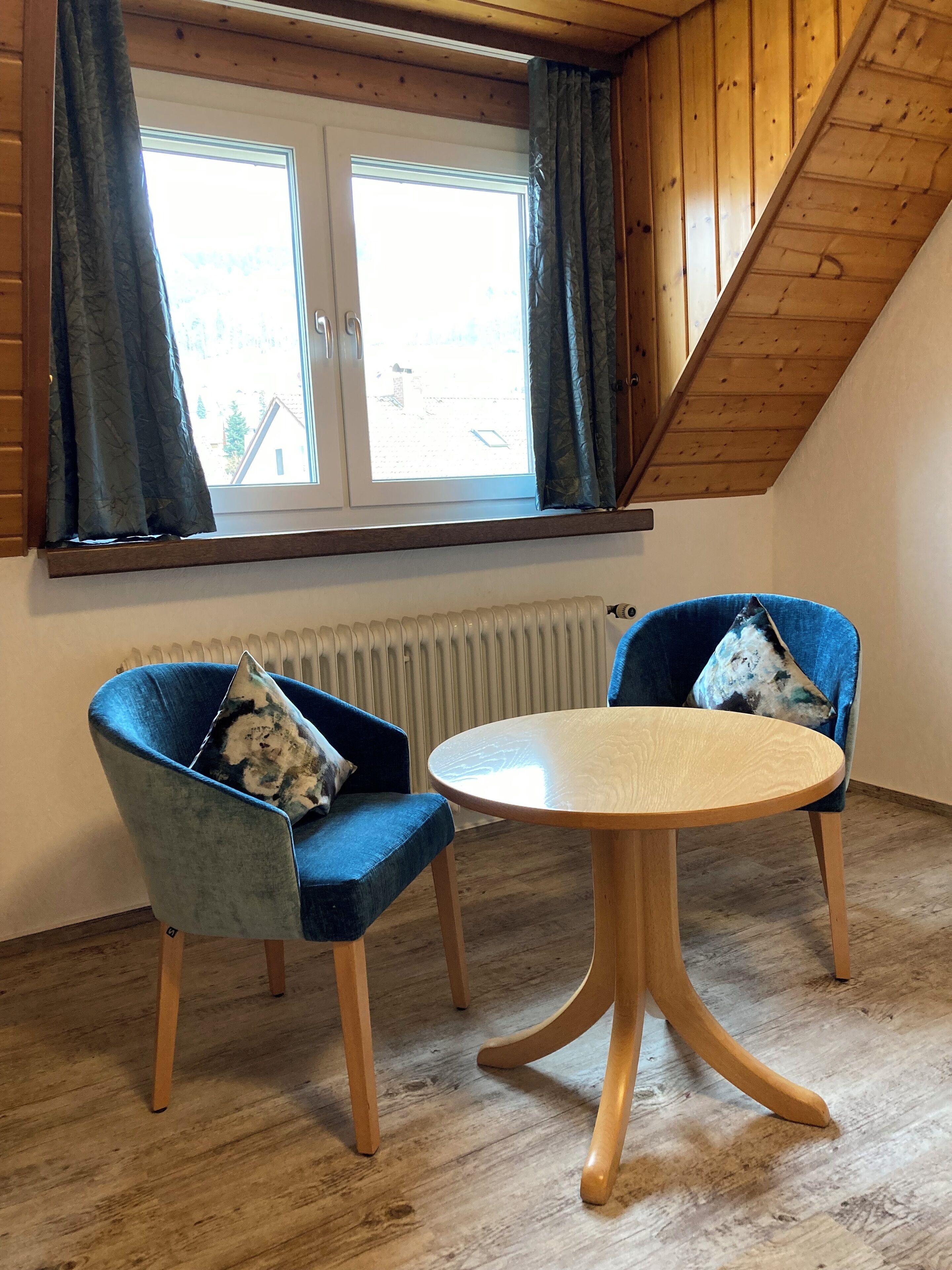 A room with two blue armchairs, a round wooden table, a window with curtains, and a sloped ceiling.