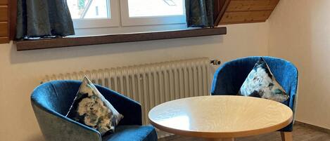A room with two blue armchairs, a round wooden table, a window with curtains, and a sloped ceiling.