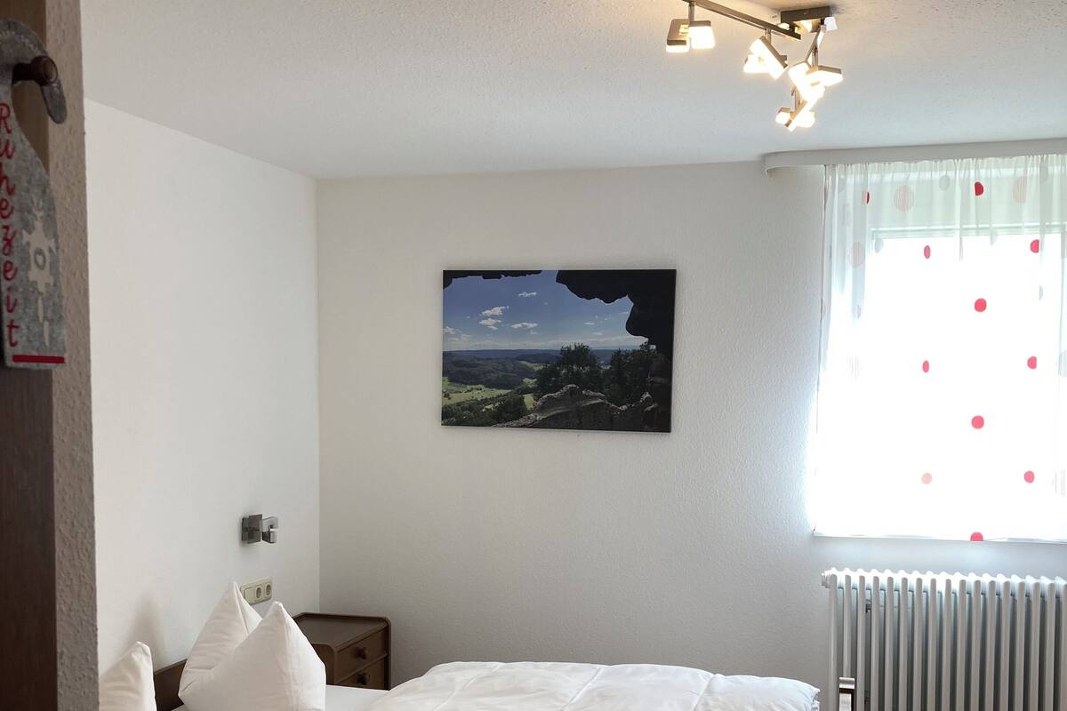 A bedroom with a bed, a nightstand, a wall-mounted light, a window with curtains, and a picture on the wall.