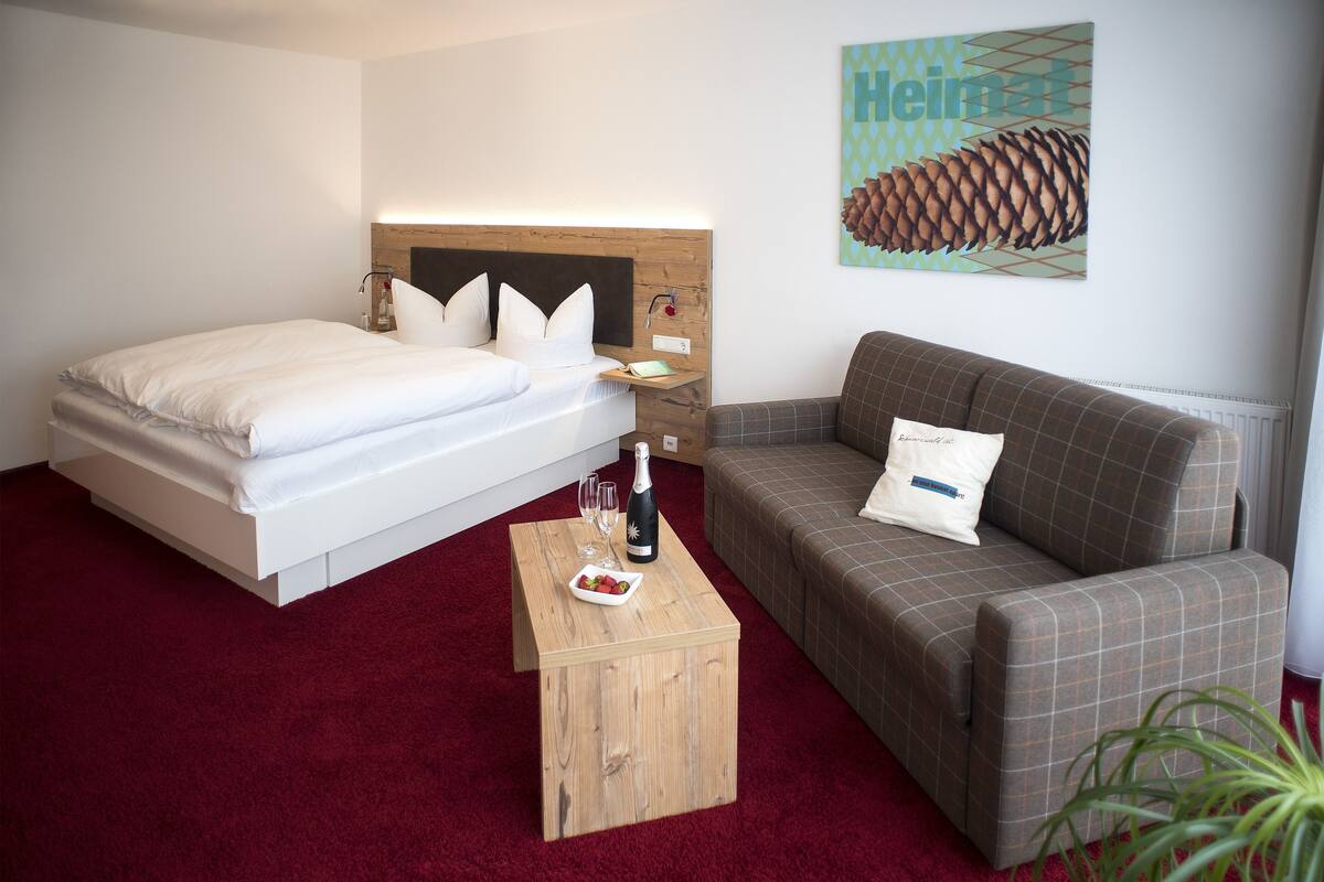 A bedroom with a bed, a wooden nightstand, a small table with a bottle and glasses, a brown sofa, and a red carpet.