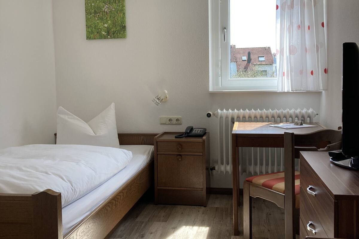 A bedroom with a bed, bedside table, desk, chair, radiator, window with curtains, and a wall-mounted light fixture.