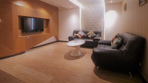 A modern living room with a flat-screen TV mounted on a wooden wall, a black leather sofa with colorful cushions, and a round coffee table. - Surreal Motel- XinJhung Branch (New Taipei City)
