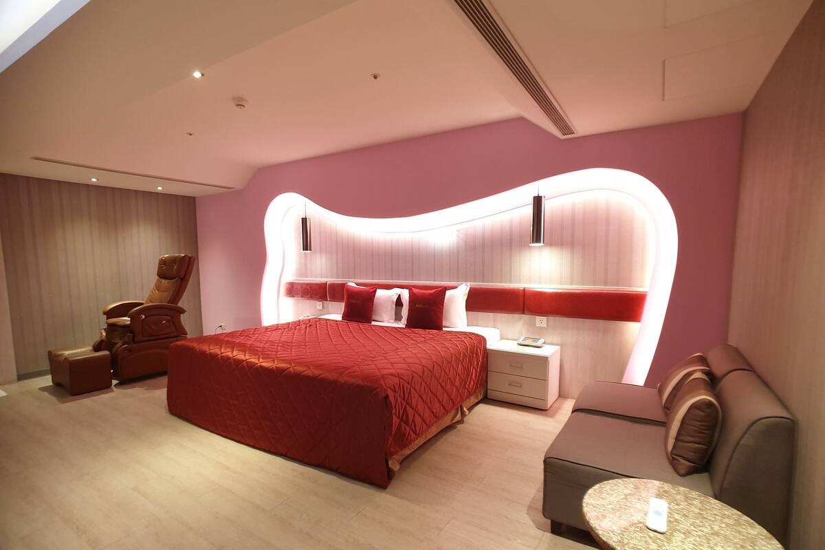 Une chambre d’hôtel moderne avec un grand lit, un fauteuil massant, une table de chevet avec une lampe et une petite table ronde.