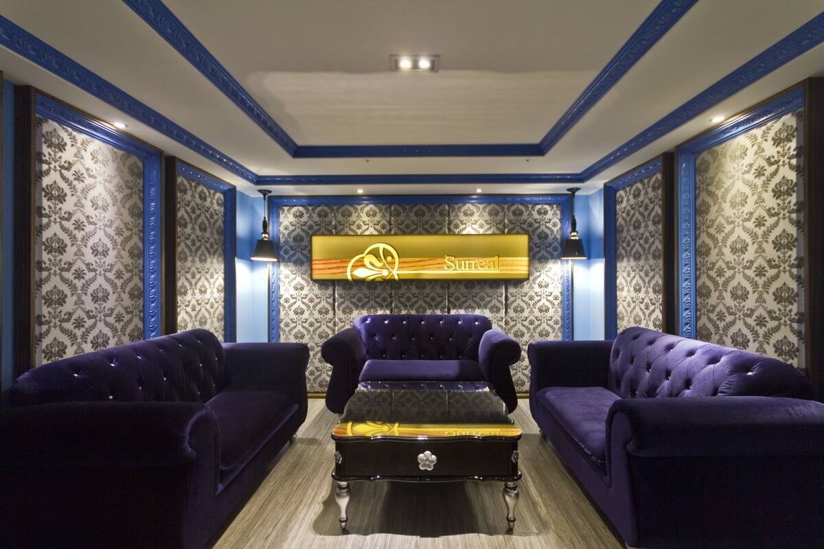 Une pièce dont les murs sont recouverts d’un papier peint à motifs bleus et blancs, avec une grande enseigne lumineuse sur laquelle on peut lire « Surréaliste », et deux canapés violets placés face à une table centrale.