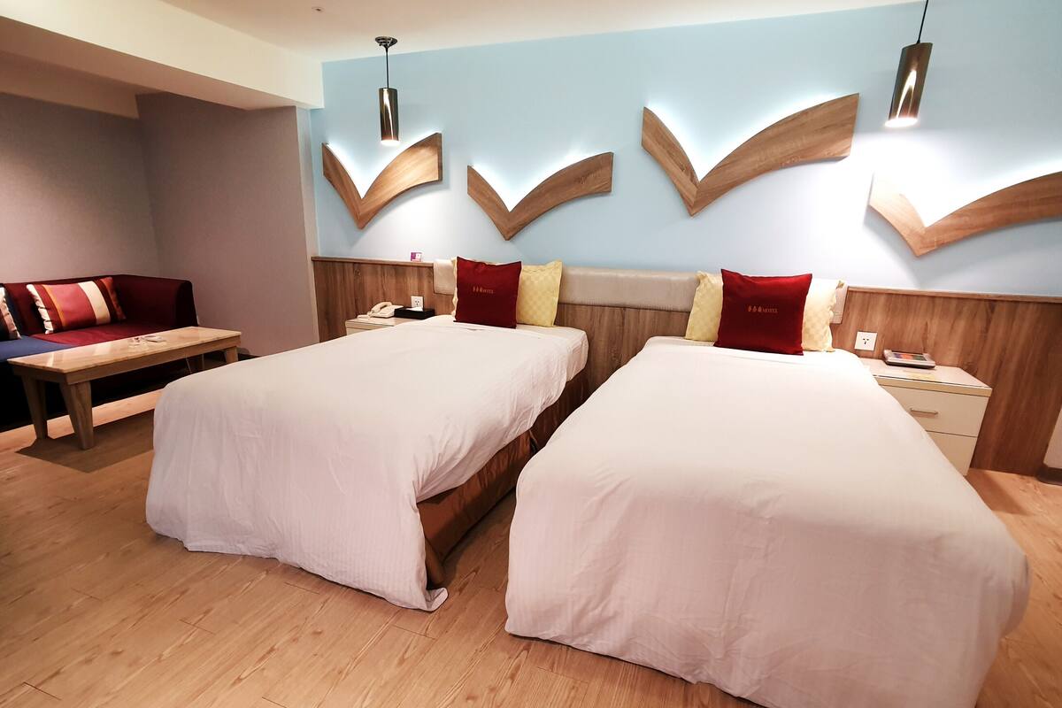 Une chambre d’hôtel avec deux lits, un banc en bois, une table de chevet et un luminaire fixé au mur.