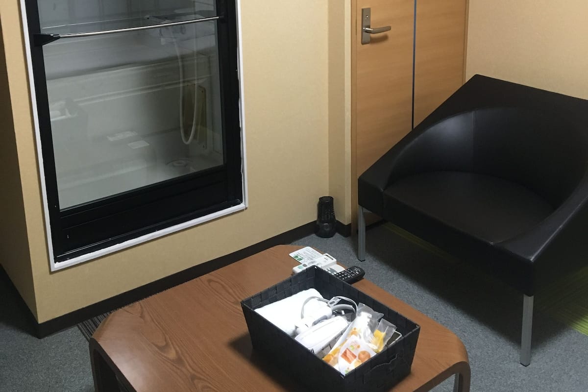 A hotel room with a wooden door, a black chair, a small table with a basket of toiletries, and a glass partition with a light fixture.