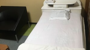 A single bed with a white mattress and a white blanket, a bedside table with a white lamp, and a black chair in a room with beige walls. - Hotel Kikuya (Kaminoyama)