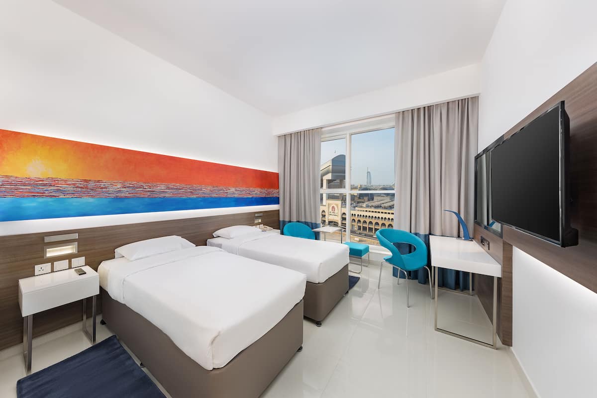 Een hotelkamer met twee bedden, een bureau, een stoel, een televisie en een groot raam met uitzicht op de stad.