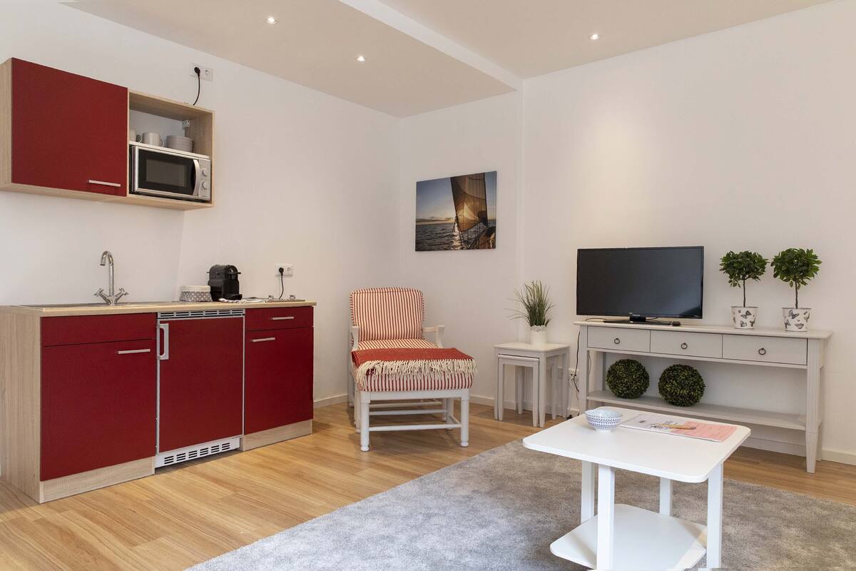 Ein modernes Wohnzimmer mit einer roten Küchenzeile, einem weißen Sessel, einem kleinen Tisch, einem Fernseher und zwei Blumentöpfen.