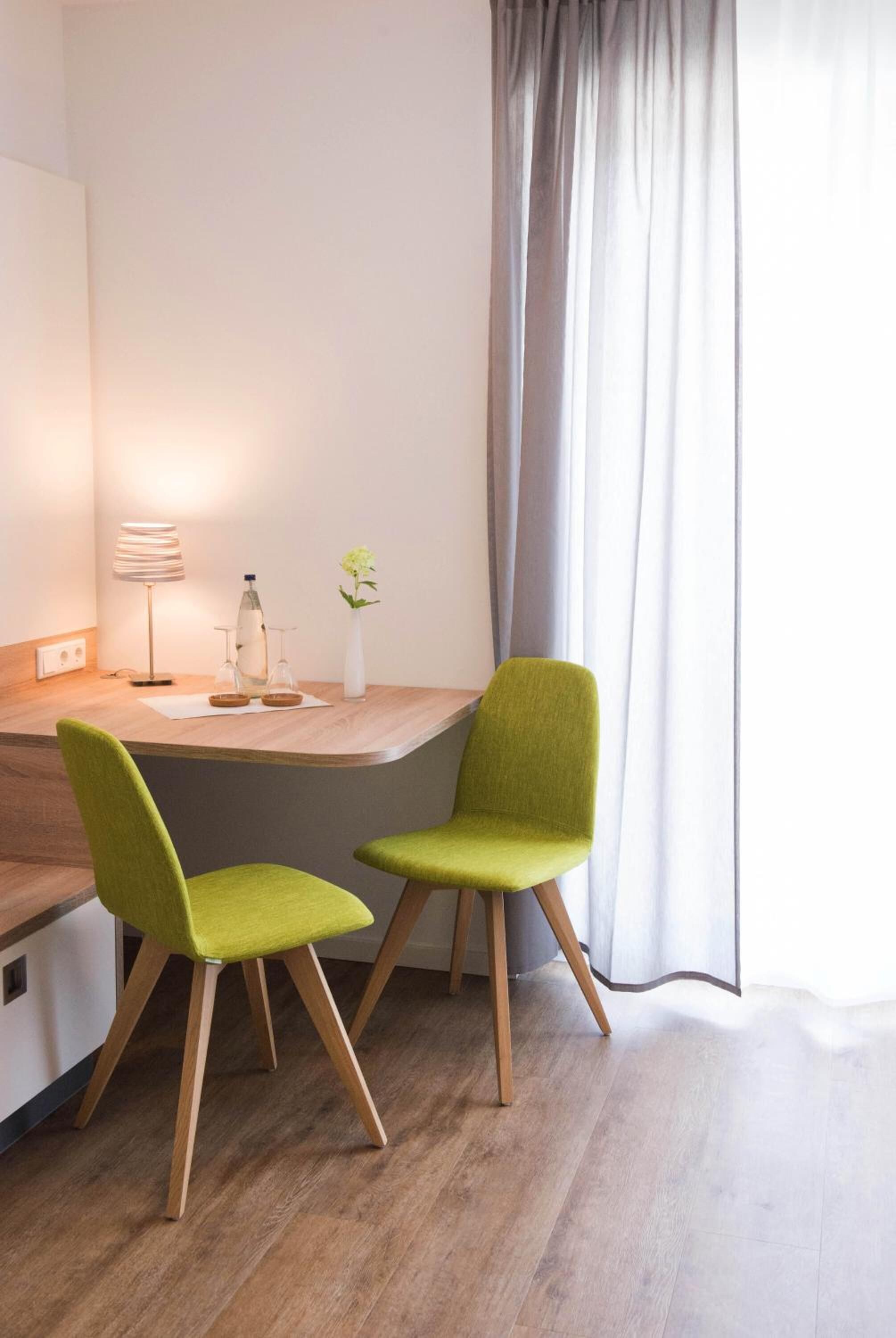 A modern room with a wooden desk, two green chairs, a lamp, a vase with flowers, and a large window with curtains.