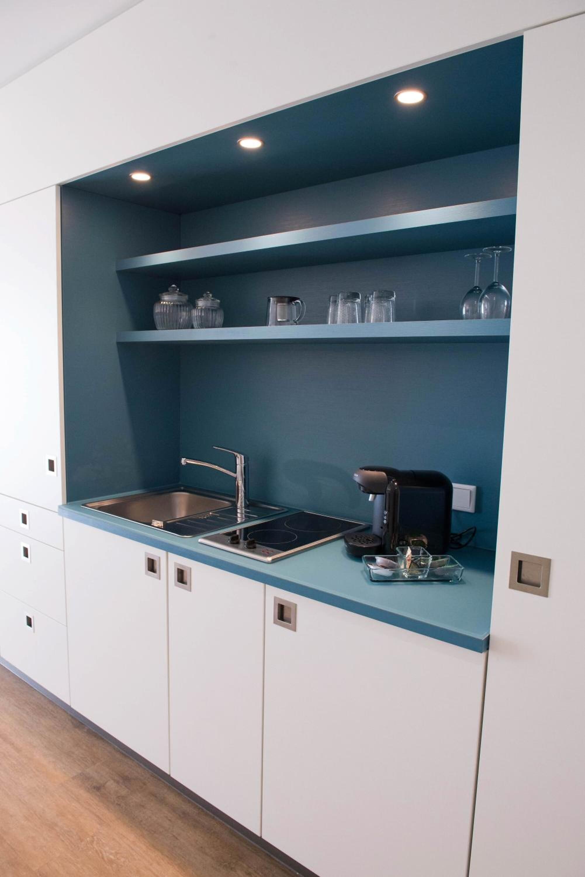 A compact kitchen with a built-in sink, a coffee maker, and shelves with glassware.