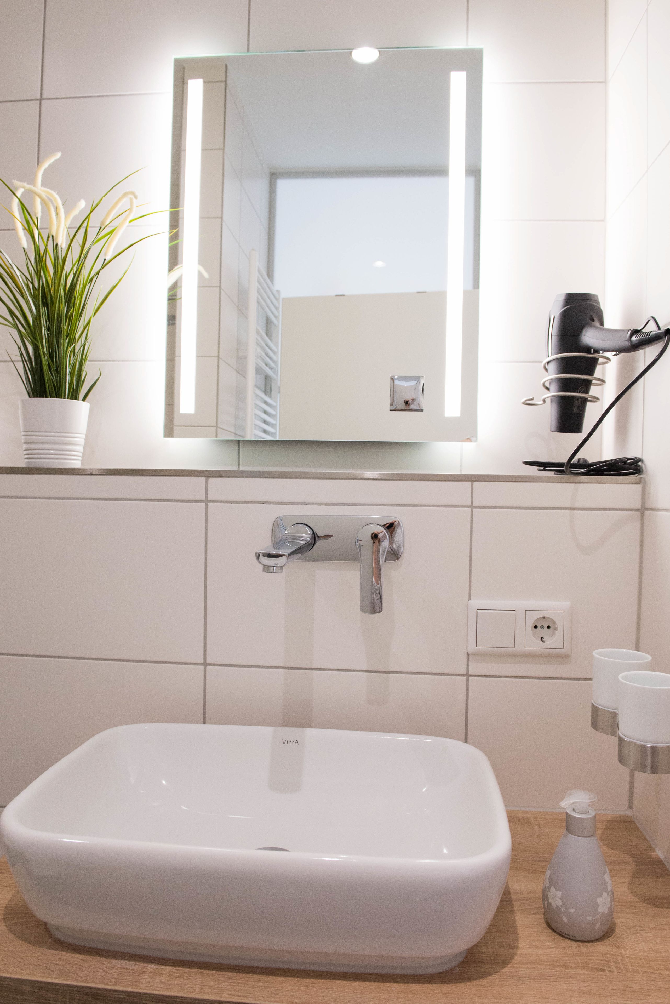 Un baño con un lavamanos blanco, un espejo, una planta en maceta, un secador de pelo y una botella de jabón para manos.
