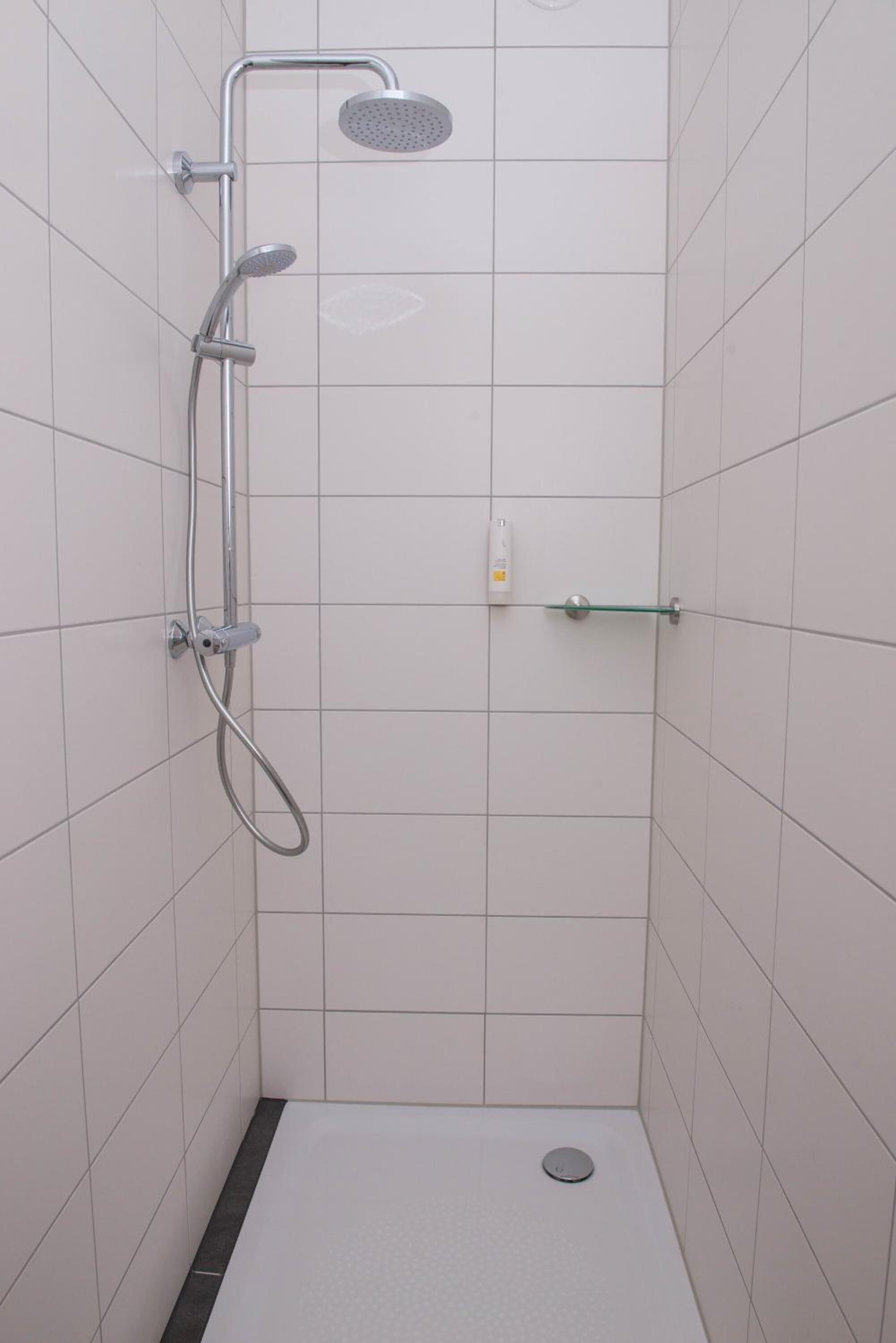 Um chuveiro pequeno com azulejos, um chuveiro manual, uma torneira fixa na parede e um prateleira de vidro.