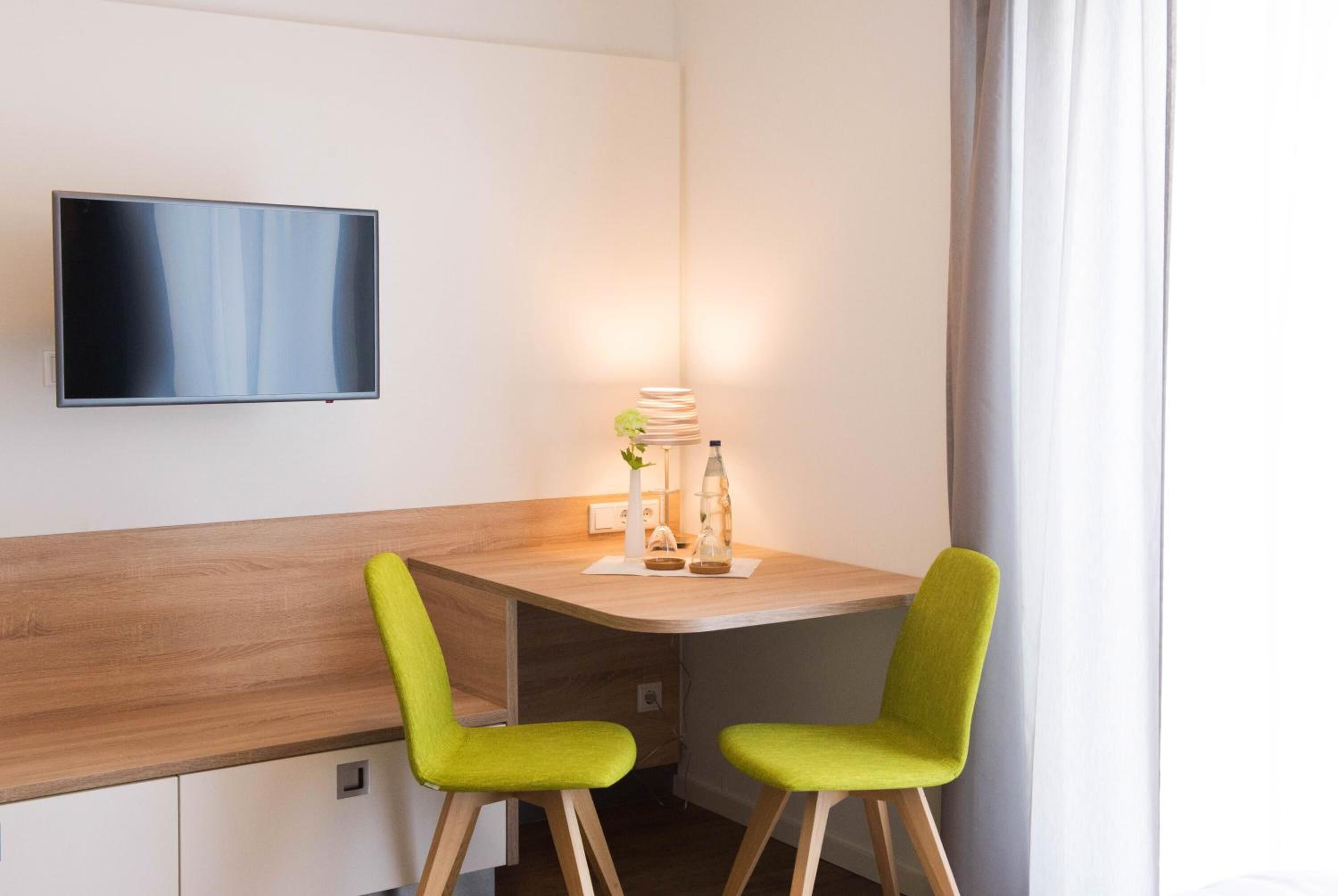 Een moderne hotelkamer met een houten bureau, twee groene stoelen, een flatscreen televisie aan de muur en een nachtkastje met een lamp en decoratieve objecten.