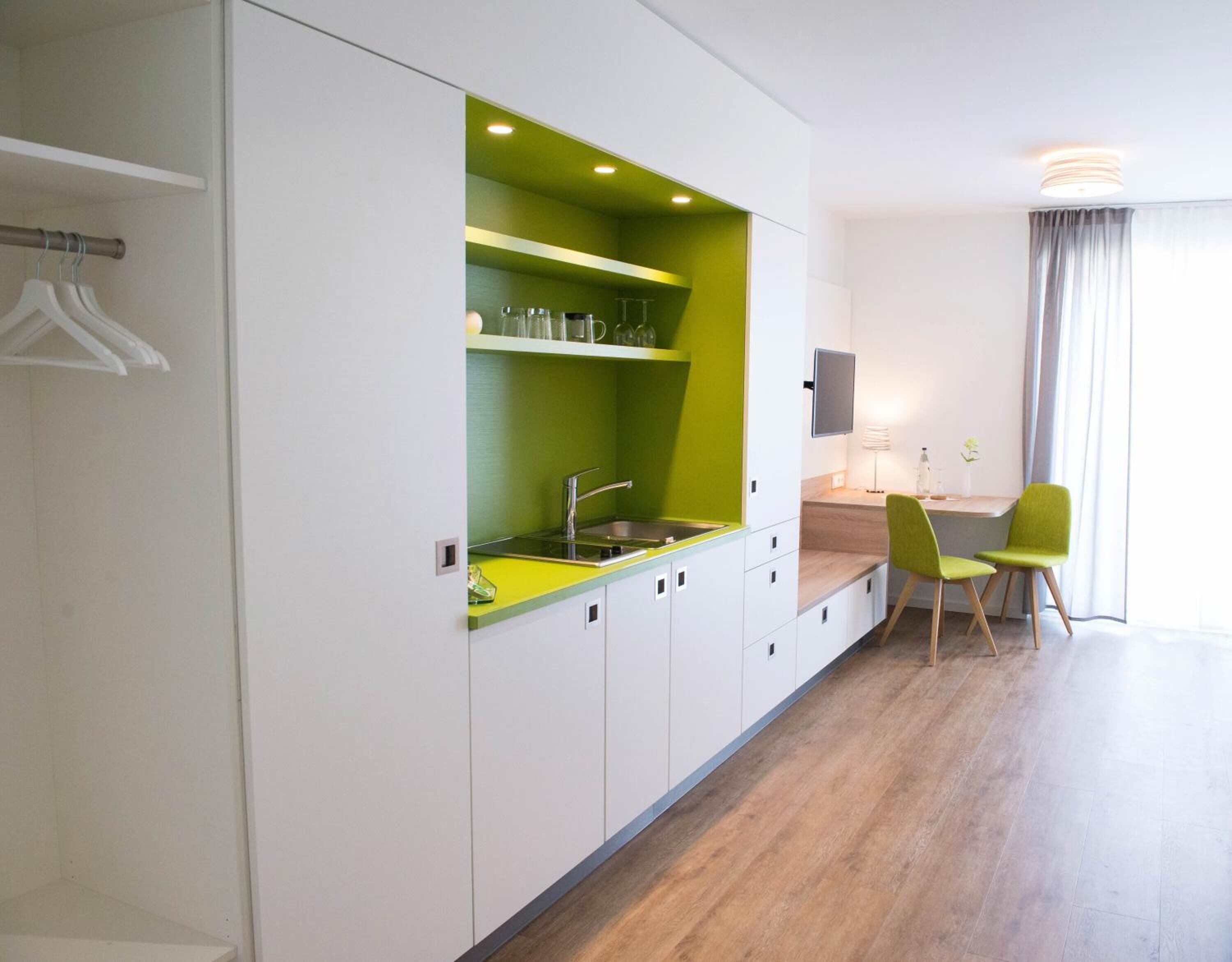 Une cuisine moderne avec des meubles blancs et un mur d’accent vert, un évier et un coin repas comprenant une table et des chaises.