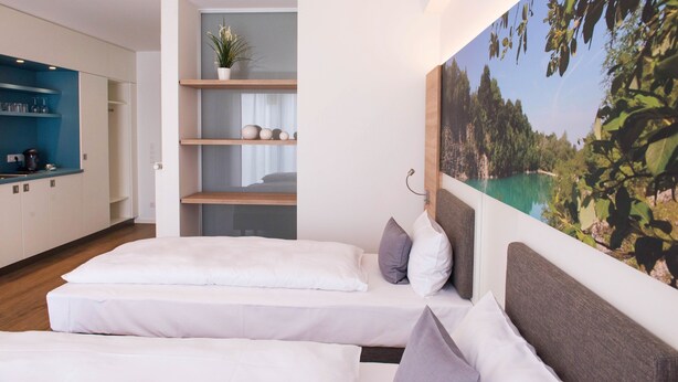 A modern hotel room with a bed, a small seating area, a kitchen, and a large window with a scenic view. - Apartment - Boardinghouse on Teuto (Lengerich)