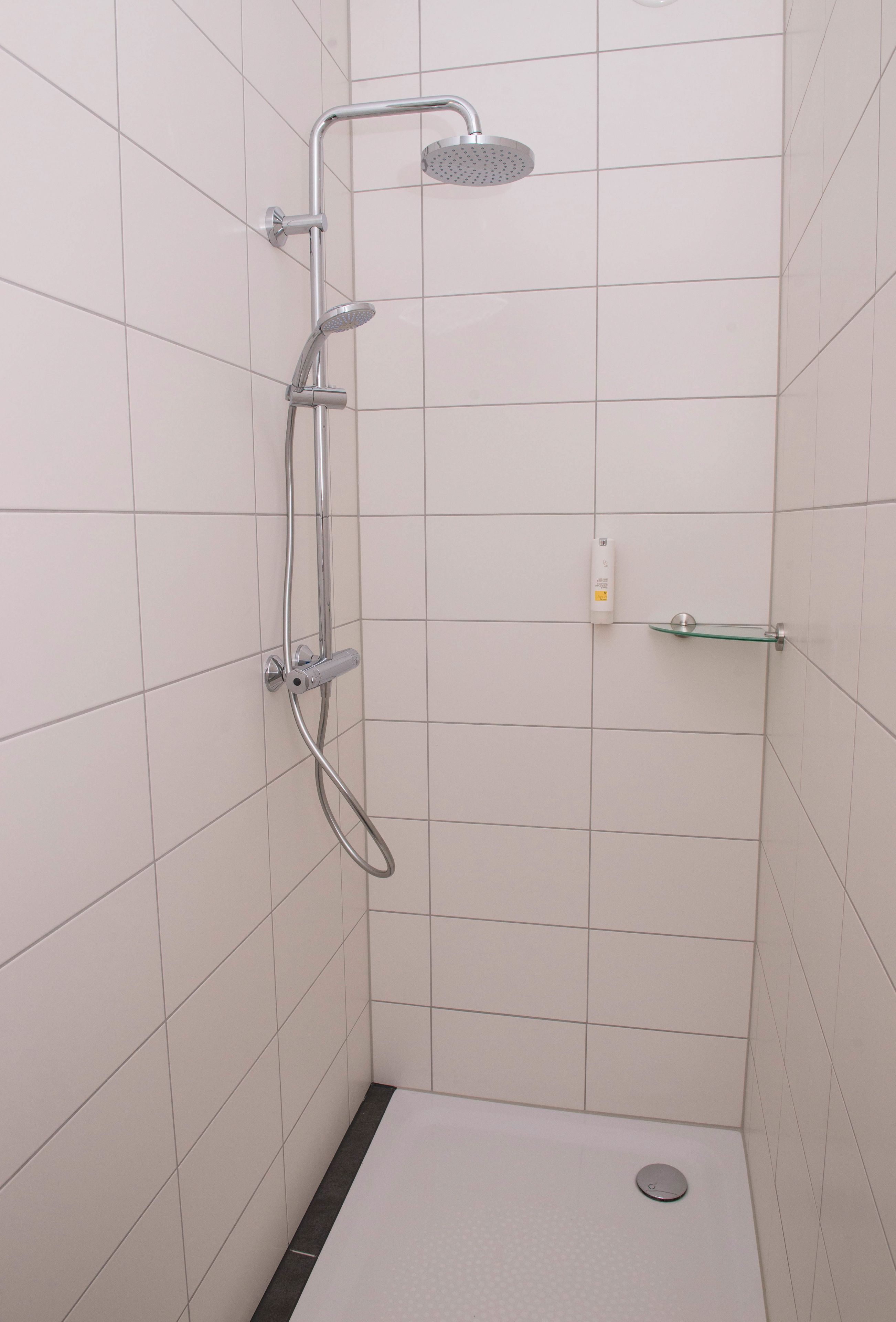 Eine kleine, mit Fliesen ausgekleidete Duschkabine mit Handbrause, Wandarmatur und Ablage aus Glas.