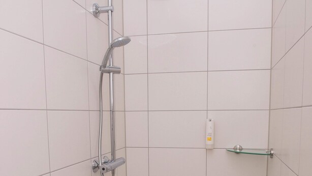 Un box de ducha pequeño con azulejos, un cabezal de ducha de mano, un grifo de pared y un estante de vidrio.