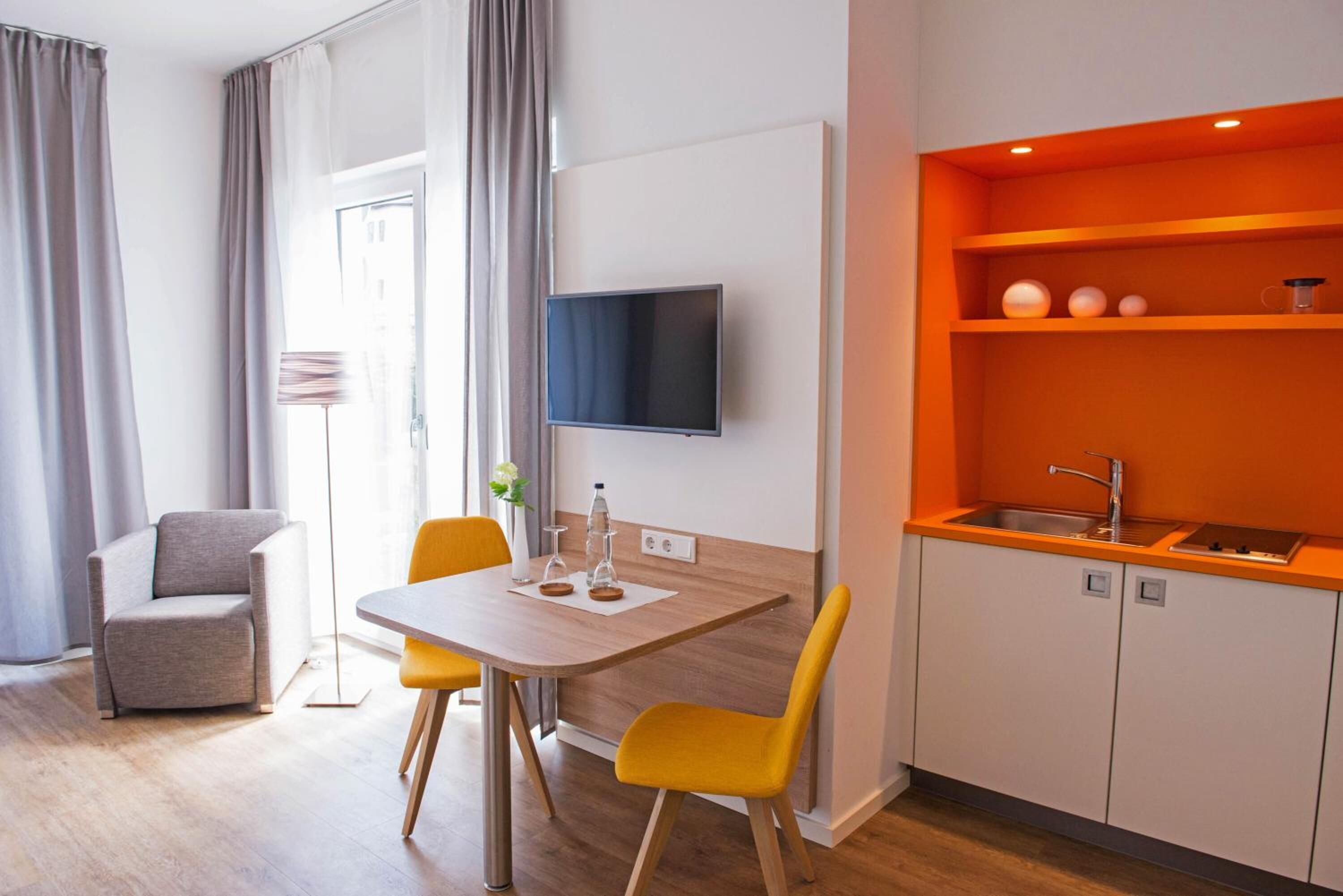 Una habitación de hotel moderna con zona de comedor, televisor de pantalla plana en la pared, cocina con estanterías color naranja y un sillón gris.