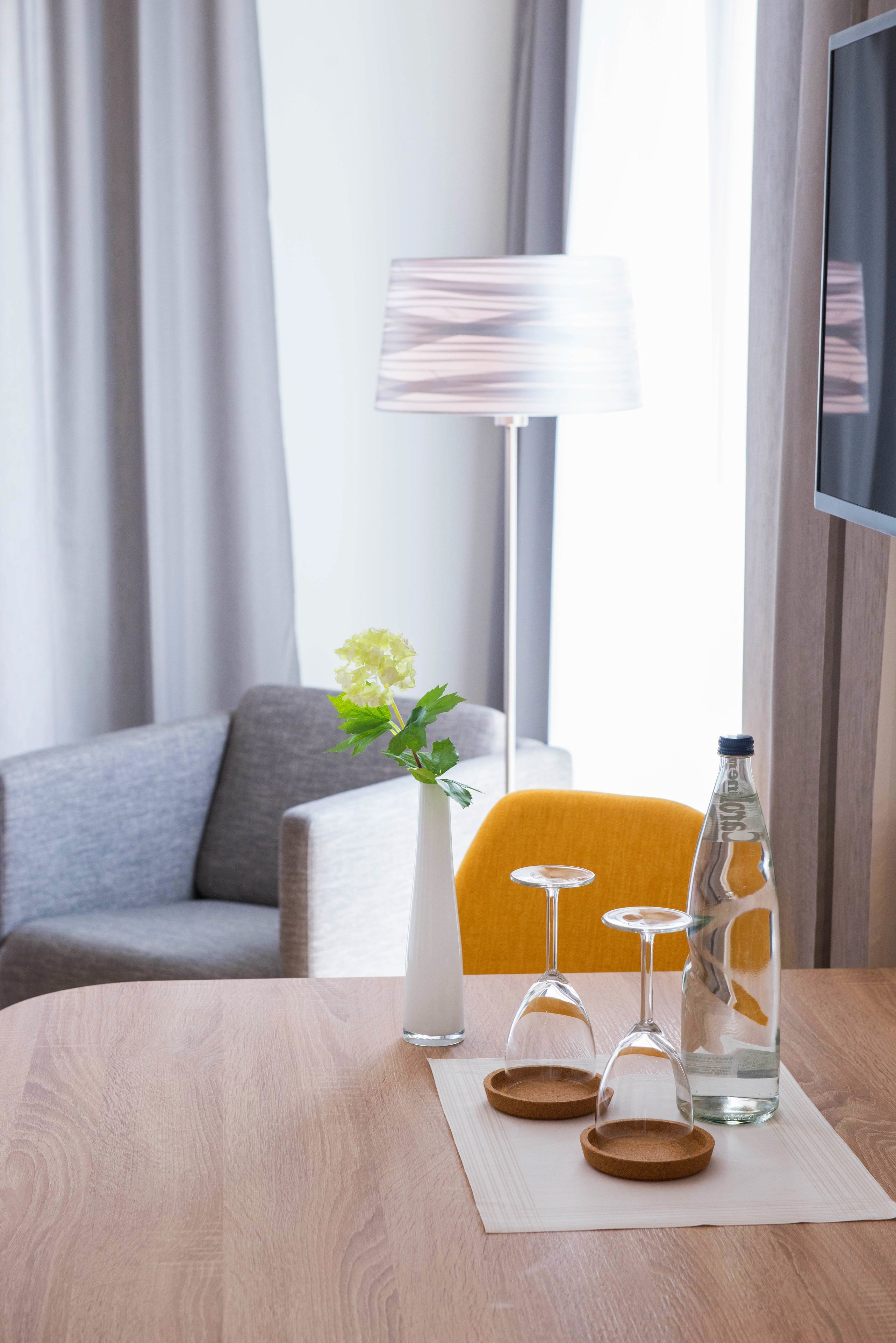 Habitación de hotel con una mesa de madera, un florero con una flor, dos copas de vino y una botella de agua.