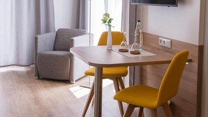 A modern hotel room with a wooden floor, a dining table with yellow chairs, a grey sofa, a flat-screen TV mounted on the wall, and a window with curtains. - Apartment - Boardinghouse on Teuto (Lengerich)
