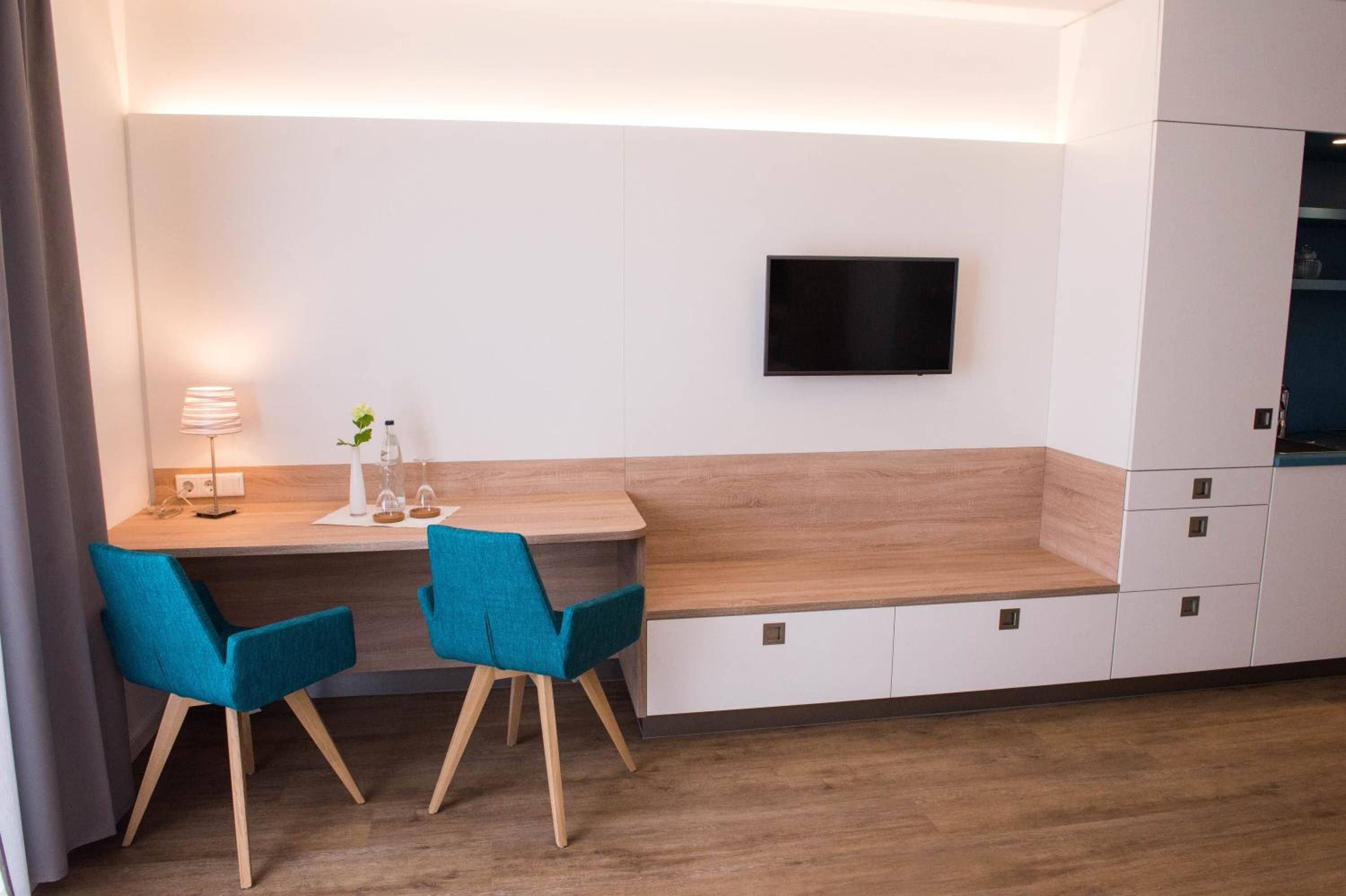 Eine moderne Küche mit integriertem Fernseher, zwei sesseln in Türkisgrün, einer Holz Arbeitsplatte und einem weißen Schrank.
