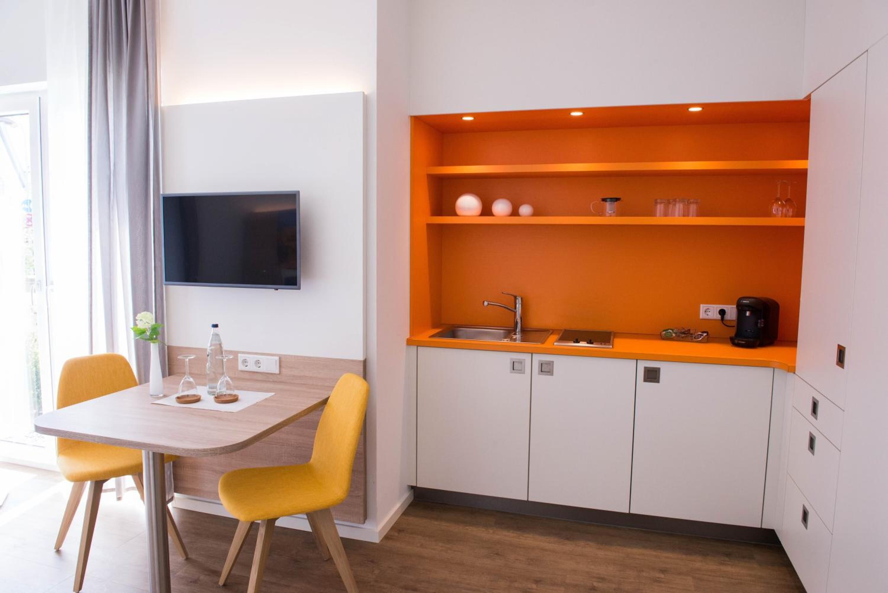 A compact kitchen with a flat-screen TV, a dining table with yellow chairs, and a bright orange accent wall.