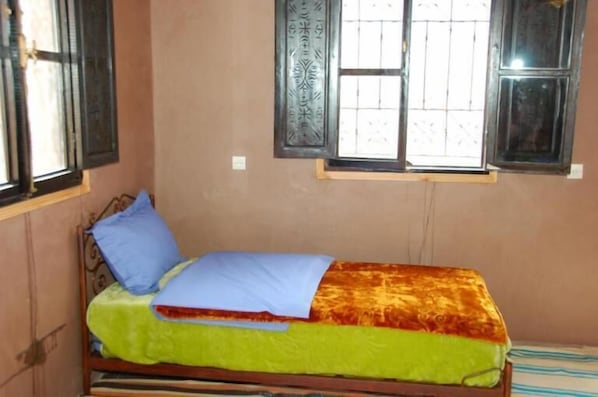 Un lit simple avec une couvre-lit verte et jaune, un oreiller bleu et une fenêtre ornée de barreaux en fer décoratifs.