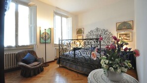 A bedroom with a metal bed frame, a rattan chair, a radiator, and a large window. - Ermione (Florence)