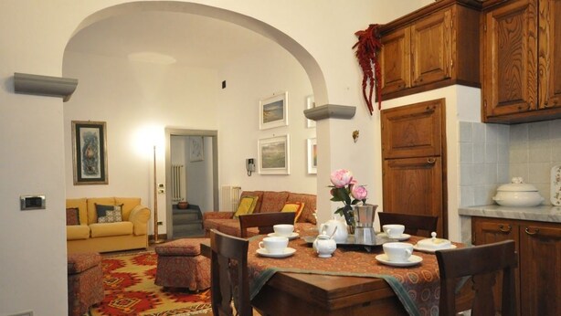A dining area with a wooden table set for four, surrounded by wooden chairs. There is a kitchen area with wooden cabinets and a countertop visible in the background. - Ermione (Florence)