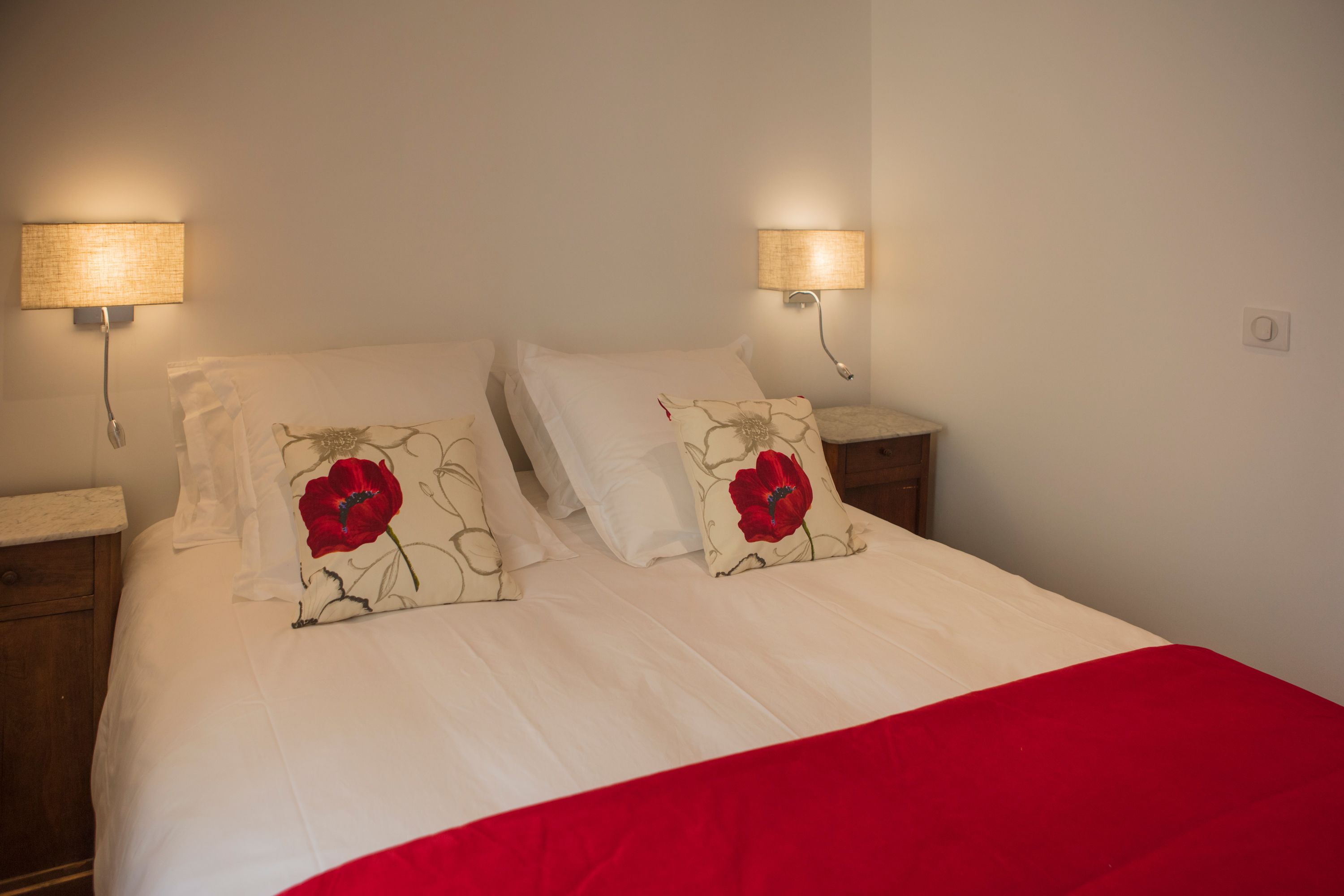 Una cama bien hecha con sábanas blancas y dos almohadas rojas con estampado floral, dos lámparas de noche y una mesita de madera.